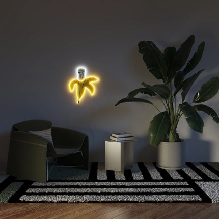 Lifestyle view of "Banana YP x JM Basquiat" neon lamp illuminated on wall in modern living room setting with chair and plant