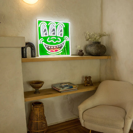 Lifestyle view of "Triple Eyes YP x Keith Haring" neon artwork displayed on a wooden shelf in a cozy room setting